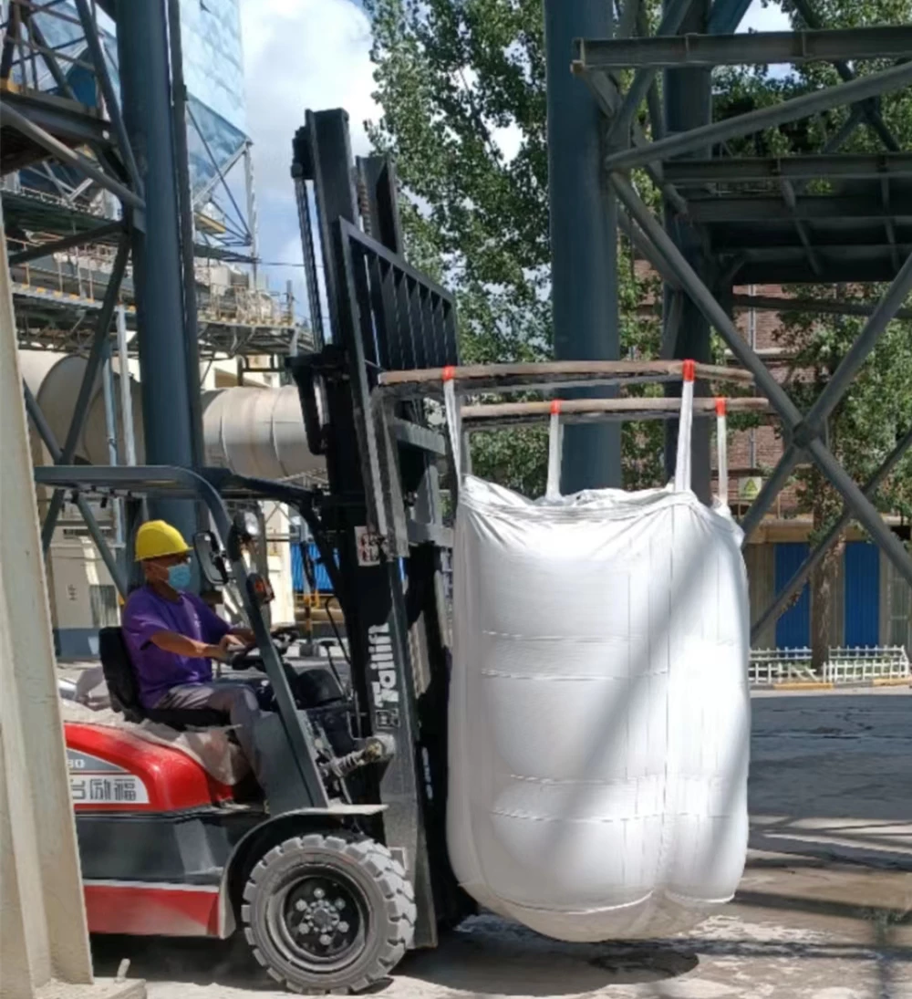 White bulk bag suspended from forklift forks by long white lifting loops in an outdoor industrial area.