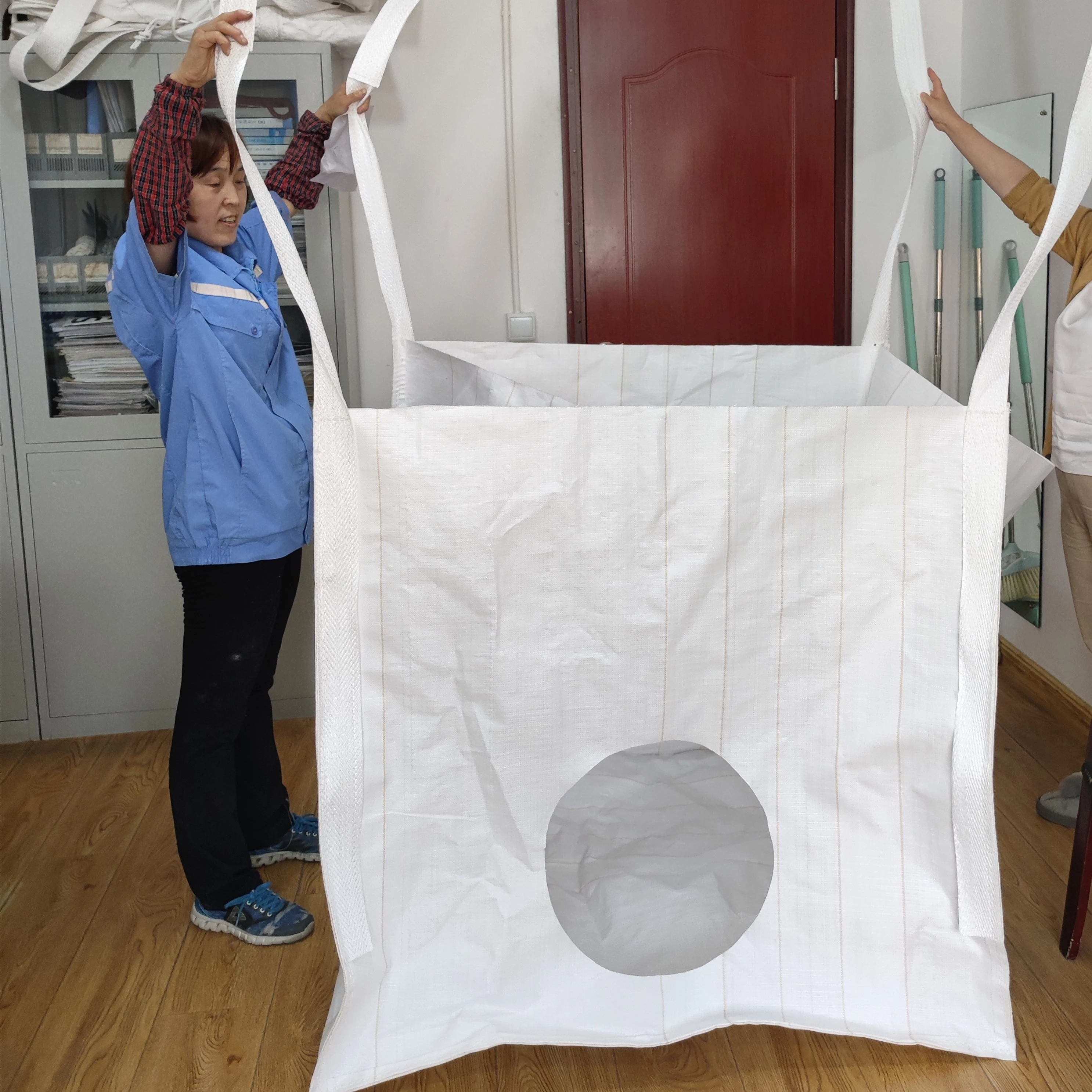 People holding an open white bag assembly with long straps and a large circular opening in the facing panel.